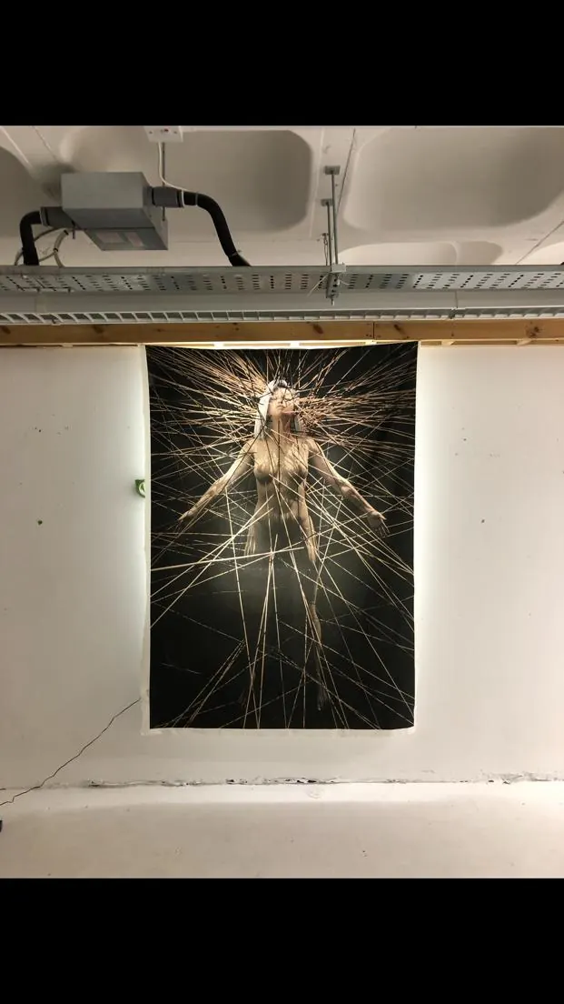 A tall dark print of a figure surrounded by radiating threads, hung in a gallery